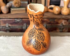A handmade gourd vase with pinecones, branches and chickadees burned into the surface of the gourd. A wavy rim has been carved in the top.